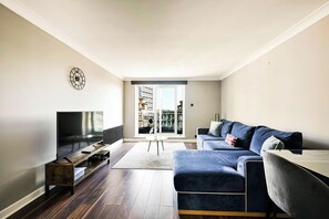 A modern living room with a flat-screen TV, a dark wood floor, a blue sofa, and a large window with a view of a building. - Maida Vale Escape (Greater London)