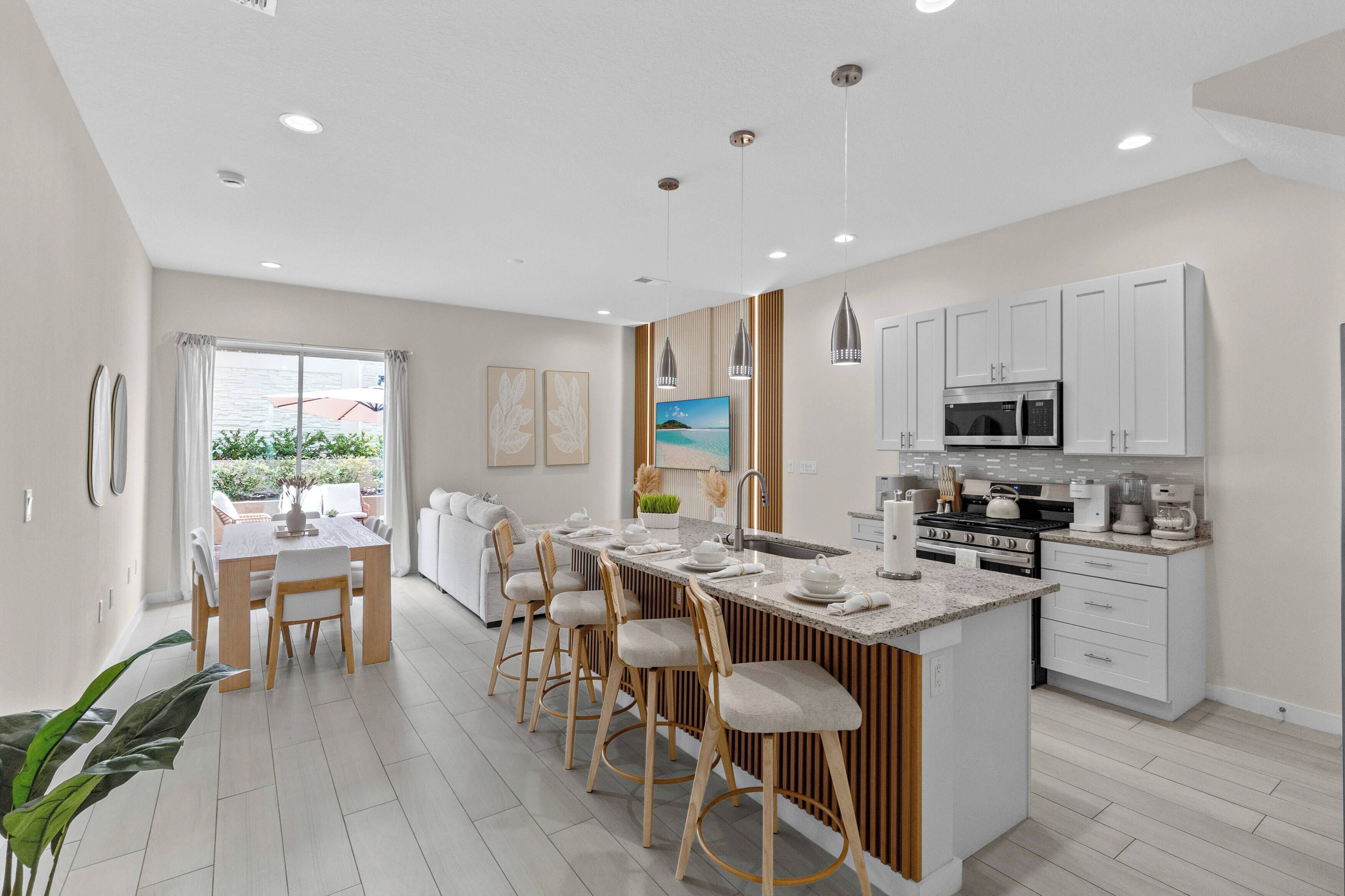 A modern kitchen with a central island, bar stools, a dining area with a table and chairs, and a view through a large window.