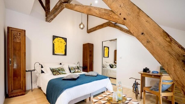 A bedroom with a sloped ceiling, a bed with a blue blanket, a wooden chair, and a table with a geometric pattern. - LA BICICLETA RAVITO (Souillac)