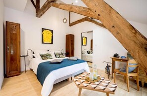 Une chambre avec un plafond en pente, un lit recouvert d’une couverture bleue, une chaise en bois et une table ornée d’un motif géométrique.