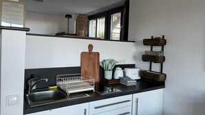 Une cuisine avec un plan de travail noir, des placards blancs, une planche à découper en bois, un égouttoir, une bouilloire, un grille-pain et une étagère en bois fixée au mur.