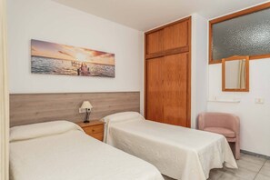 Ein Schlafzimmer mit zwei Betten, einem Holzschrank, einem Nachttisch mit Lampe, einem rosa Sessel und einem Gemälde, das einen Sonnenuntergang über Wasser zeigt.