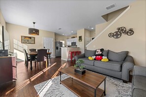 Ein modernes Wohnzimmer mit grauem Sofa, einem Essbereich mit Holztisch und einer Küche im Hintergrund.