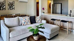 Una sala de estar moderna con un sofá, una mesa de centro con una planta, un ventilador de techo y una zona de cocina con un refrigerador y un lavamanos.