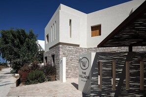 A modern building with a white facade, a stone base, and a sign reading "The Village Milos". - The Village by WPS (Milos)