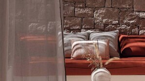 A room with a stone wall, a round white table with a vase, and a red sofa with patterned cushions. - The Village by WPS (Milos)