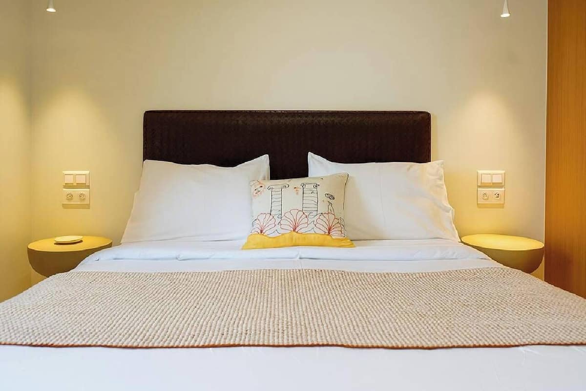 Una cama bien hecha con sábanas blancas y un cojín decorativo, flanqueada por dos mesitas de noche con lámparas, en una habitación bien iluminada.