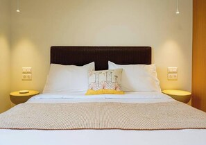A neatly made bed with white linens and a decorative pillow, flanked by two bedside tables with lamps, in a well-lit room. - The Village by WPS (Milos)