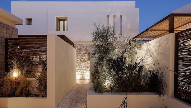 A modern building with a clean, white facade, a stone wall, and a small garden area with plants and a pathway. - The Village by WPS (Milos)