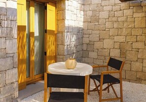 A stone wall, a wooden table with a vase, and two chairs in a courtyard. - The Village by WPS (Milos)