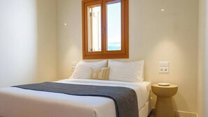 A bedroom with a bed, a bedside table, a window, and a wall socket. - The Village by WPS (Milos)