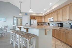 A modern kitchen with wooden cabinets, a white island with bar stools, and a dining area with white chairs. - Sunset Bay Villa 320 by Seaside Vacations and Sales (Chincoteague Island)