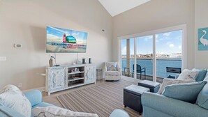 Un soggiorno spazioso con una grande televisione, posti a sedere confortevoli, vista sul mare e un balcone con mobili da esterno.