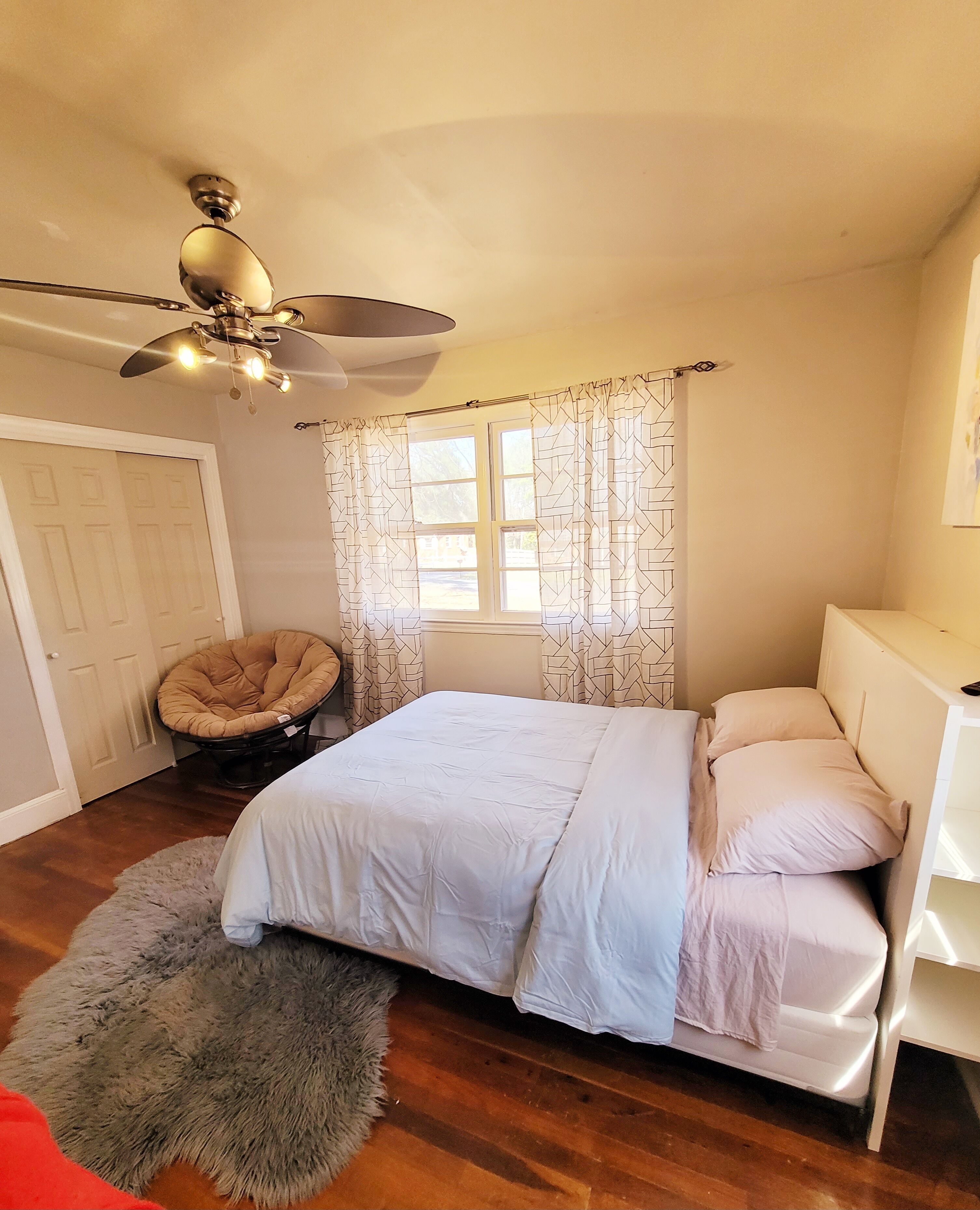 Une chambre à coucher comprenant un lit, une chaise, un ventilateur de plafond, une fenêtre avec des rideaux et une porte.