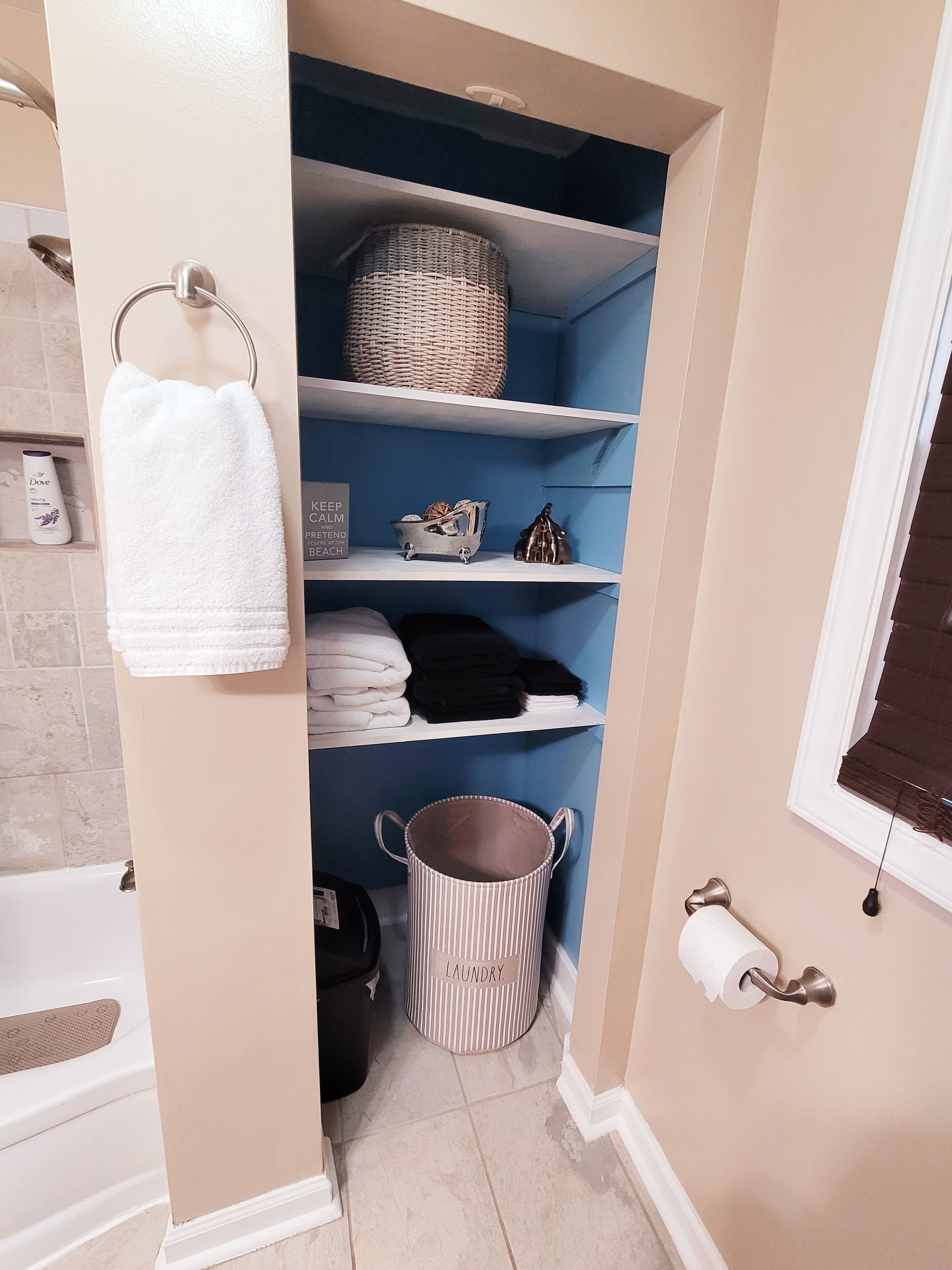 A bathroom with a white bathtub, a towel rack with a white towel, shelves with folded towels and toiletries, a striped laundry basket, and a toilet paper holder.