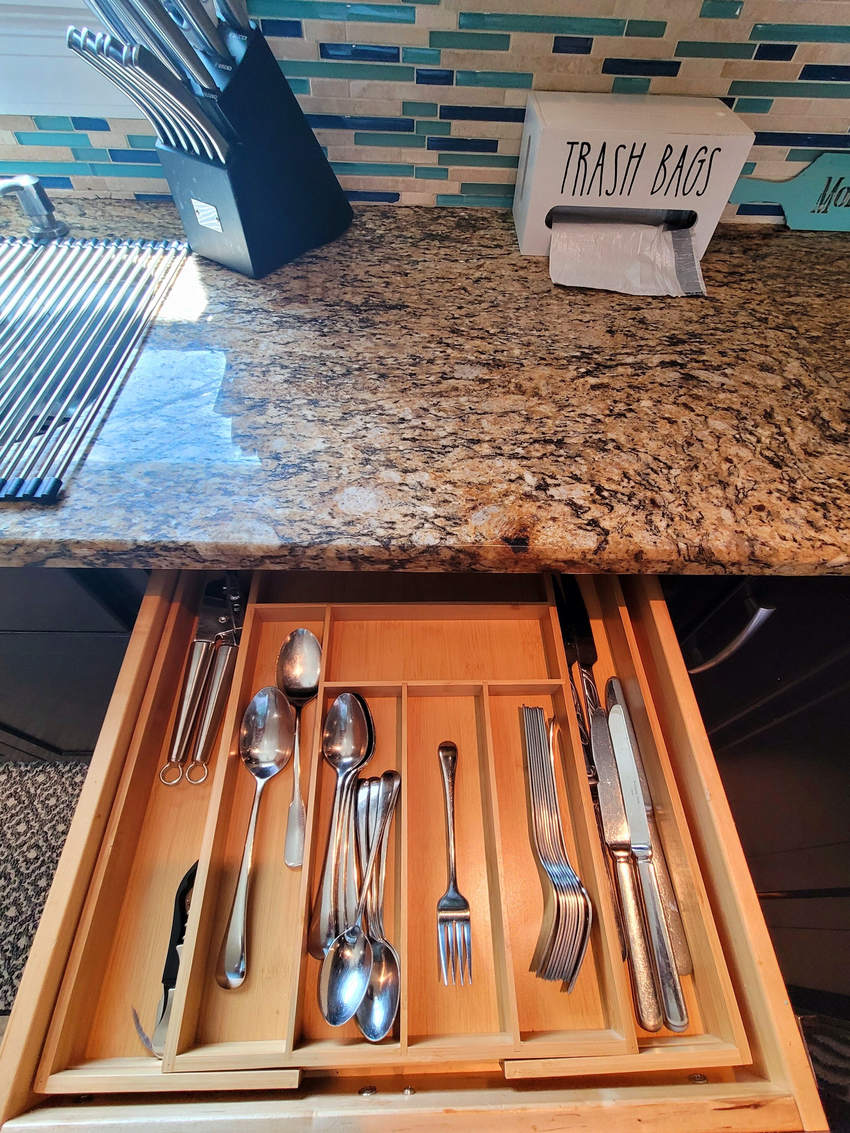 Un cajón de cocina con utensilios organizados, un dispensador de bolsas de basura en la encimera y un salpicadero con baldosas azules y beige.