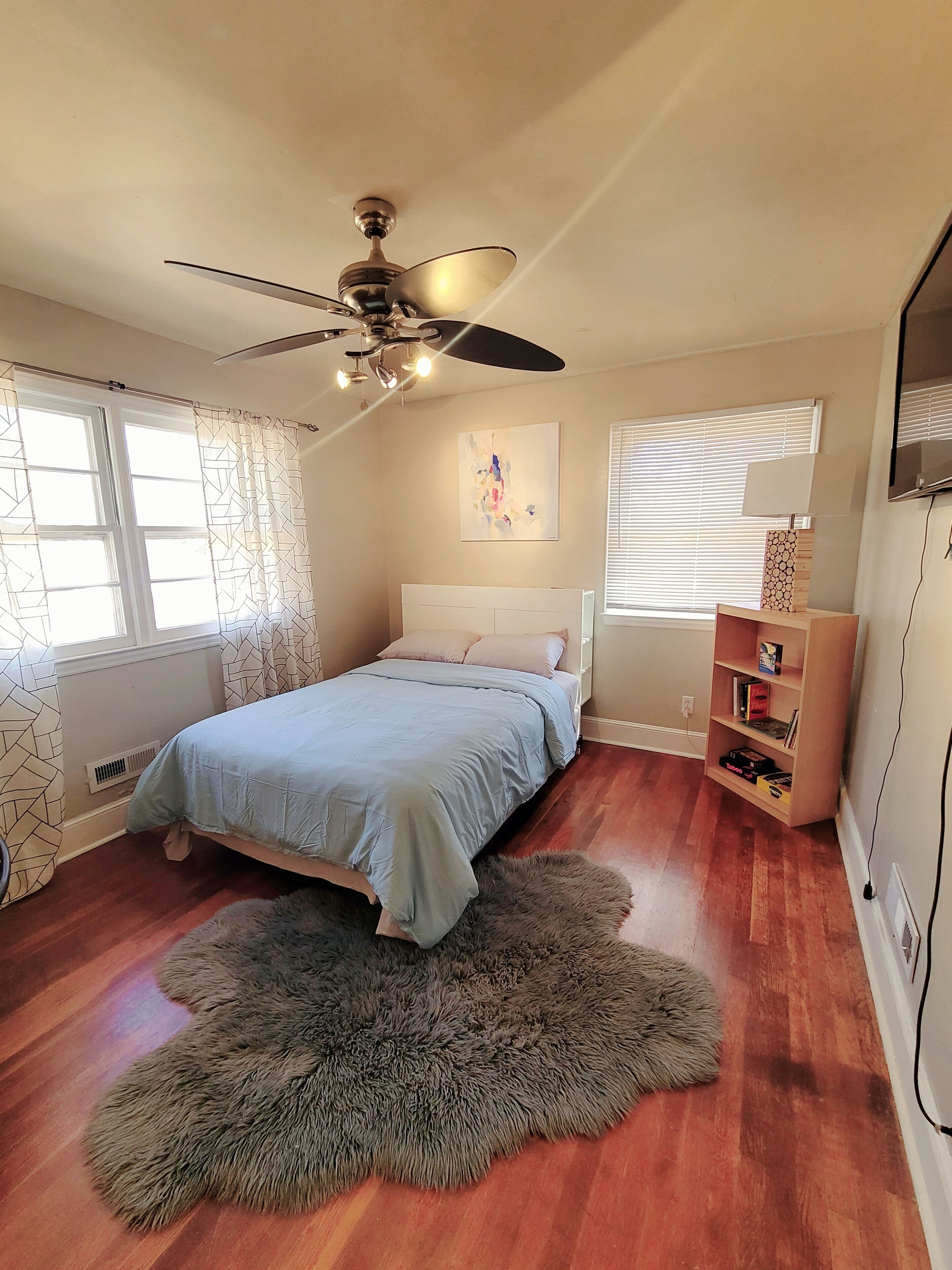 A bedroom with a bed, a ceiling fan, a window with blinds, a bookshelf, and a rug.