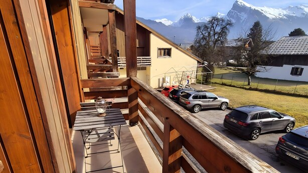 Un balcón de madera con vistas a montañas nevadas, un aparcamiento cubierto con coches estacionados y un edificio con techo inclinado.