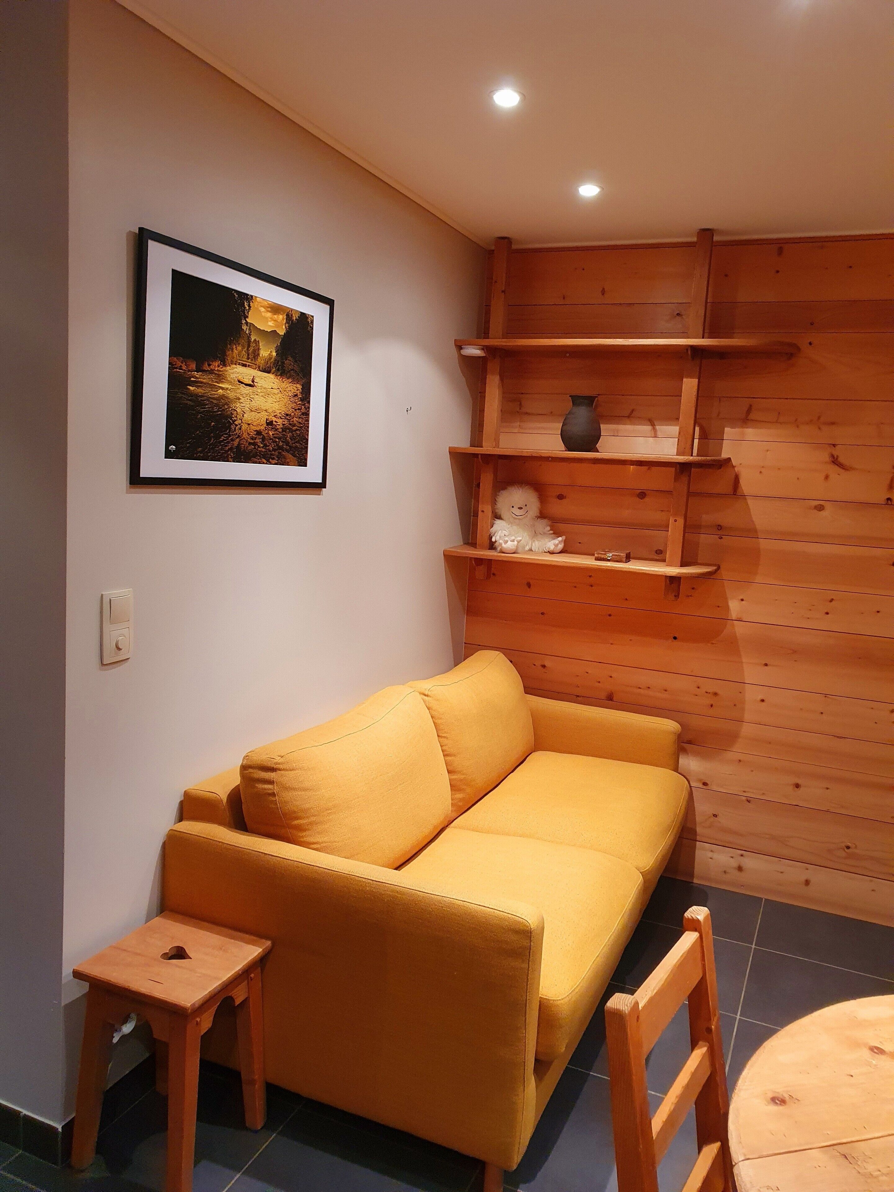 A yellow sofa, a small wooden stool, a wooden shelf unit, and a framed picture on the wall.