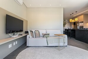 A modern living room with a white sofa, a flat-screen TV, and a small wooden side table. - Wi-Fi 400mb Smart TV 50 inches Beach 1.5 km #CHA13 (Florianópolis)