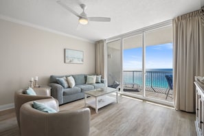 Ein geräumiges Wohnzimmer mit grauem Sofa, weißem Couchtisch und beigefarbenem Sessel. Ein Deckenventilator ist sichtbar, und das Zimmer verfügt über eine große Schiebeglasdoor zum Balkon mit Meerblick.