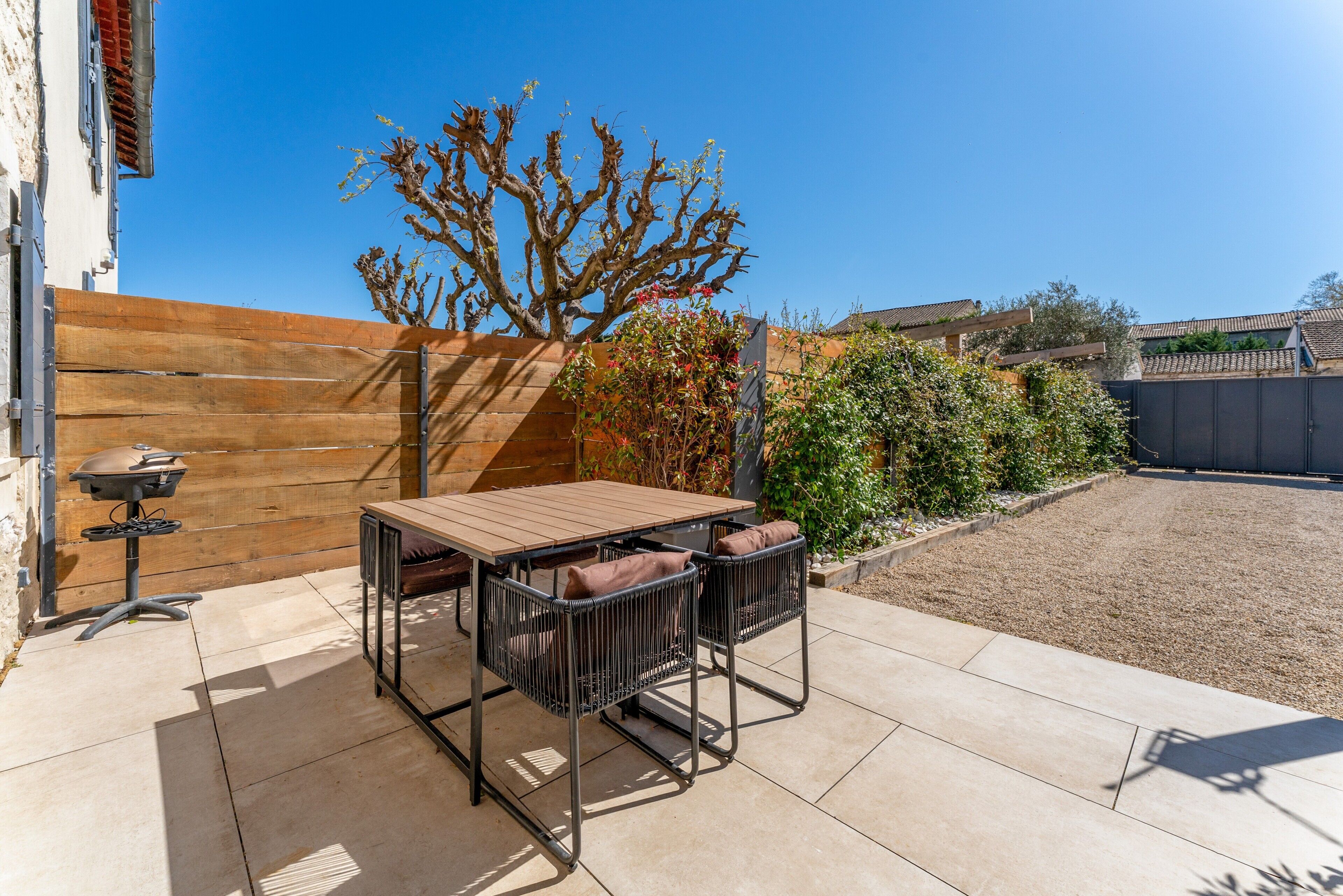 Een terras met een houten tafel en stoelen, een barbecue en uitzicht op een tuin met een boom en struiken.