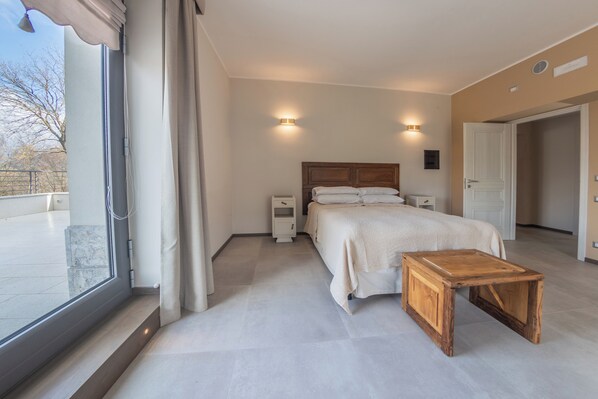 Un dormitorio con una cama grande, mesitas de noche, una mesita auxiliar de madera, una ventana con cortinas y vistas al exterior.