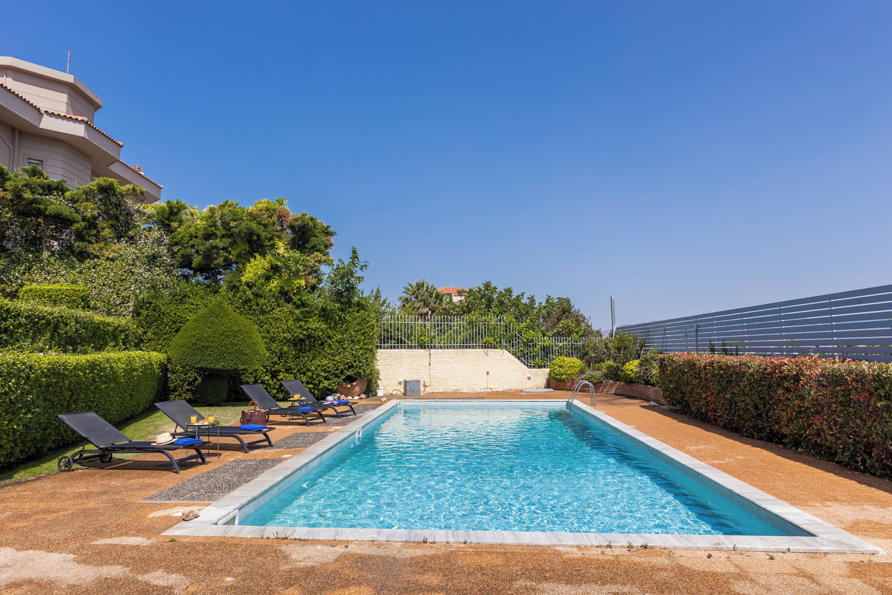 A rectangular swimming pool with a clear blue water surface, surrounded by a low brick wall. There are several lounge chairs with blue cushions and a small table with a bottle and a glass. The pool area is adjacent to a well-maintained hedge and a building with a flat roof.