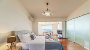 A modern bedroom with a bed, bedside table, and a view of the sea through a large window. - Blue Horizon Villa (Kalyvia Thorikou)