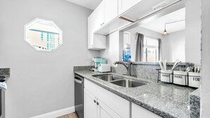 A modern kitchen with white cabinets, granite countertops, a double sink, and a view of a building through a hexagonal window. - 🏖️Easy Beach Access | Family Friendly | Sand Castles West II G3- Castawave🏖️ (Panama City Beach)