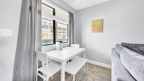 A modern room with a white table and chairs, a grey sofa, and a window with blinds. - 🏖️Easy Beach Access | Family Friendly | Sand Castles West II G3- Castawave🏖️ (Panama City Beach)