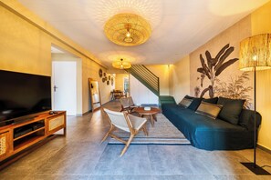 Ein modernes Wohnzimmer mit Flachbildfernseher, Holzunterhaltungseinheit, dunkelblauem Sofa, Korbsesseln, einem runden Couchtisch und einer Treppe, die zu einer weiteren Ebene führt.