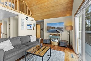 A modern living room with a wooden ceiling, a grey sofa, a coffee table, and a large window with a view of a lake. - True Lakefront/Private Dock/15 mins to Crosslake (Fifty Lakes)
