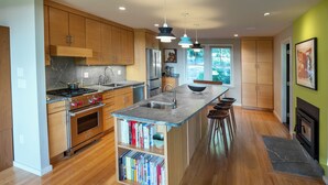 A modern kitchen with wooden cabinets, a central island with stools, a built-in bookshelf, and a large window providing natural light. - 1 of a Kind: Modern Architecture, Natural Beauty, Walk to Amherst College (Amherst)