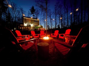 A cozy outdoor seating area with red Adirondack chairs around a central fire pit, set against a backdrop of trees and a house. - Luxe North Conway Family Retreat -Sleeps 12- Hot Tub + Game&Movie Room+Kids Loft (Conway)