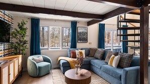 A cozy living room with a blue sofa, a wooden coffee table, and a blue armchair. There is a window with curtains, a bookshelf, and a plant. - Luxe North Conway Family Retreat.Sleeps 12.Hot Tub+Game&Movie Rm+Kids Loft+Pets (Conway)