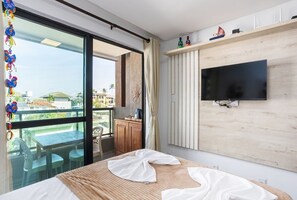 A hotel room with a bed, a TV mounted on the wall, a balcony with a table and chairs, and a view of the cityscape. - Villa Del Mar - Studios in Porto de Galinhas by Carpediem (Ipojuca)