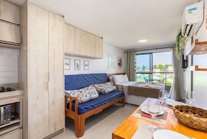 A compact living space with a kitchenette, a sofa with floral cushions, a dining table with wine glasses, and a view of the outdoors. - Villa Del Mar - Studios in Porto de Galinhas by Carpediem (Ipojuca)