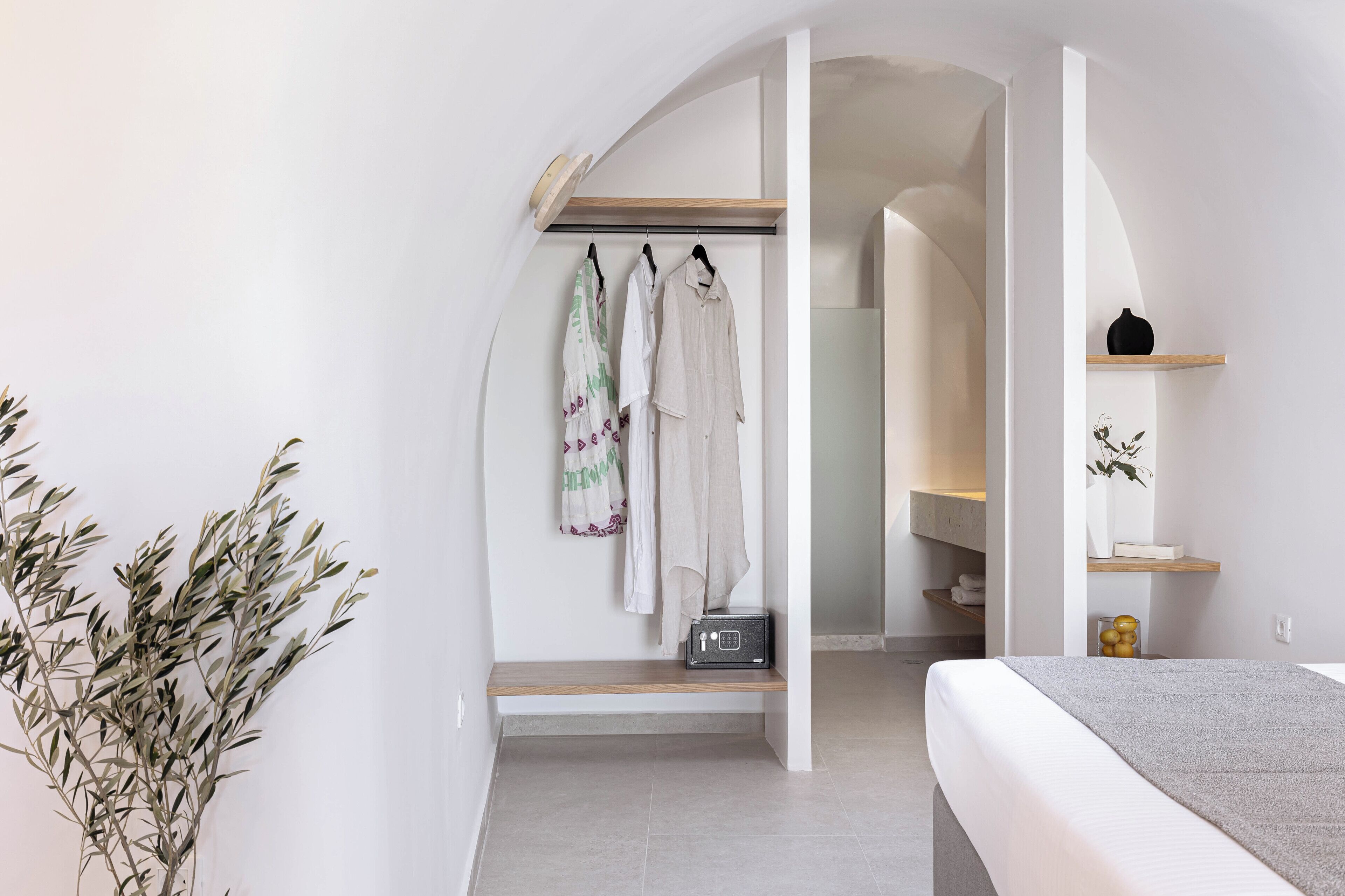 A modern bedroom with a bed, a bedside table, a shelf with decorative items, and a walk-in closet with hanging clothes and a radio.