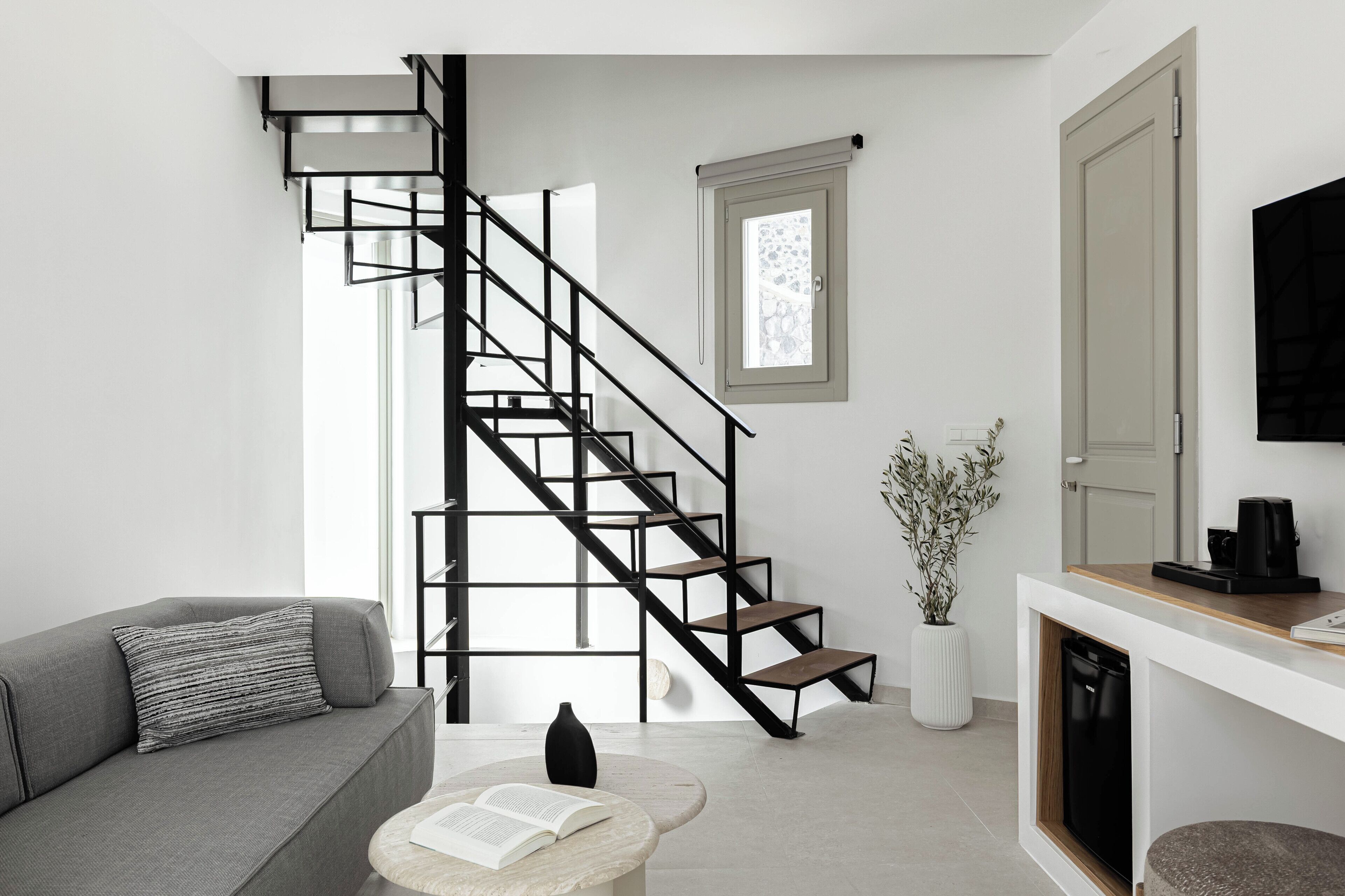 A modern living room with a grey sofa, a round coffee table with a book, a black vase, a staircase with black railings, a white door, and a potted plant.