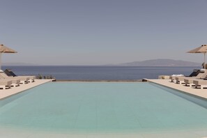 A rectangular swimming pool with lounge chairs and umbrellas on both sides, overlooking a body of water and distant land. - Filomare Suites (Santorini)