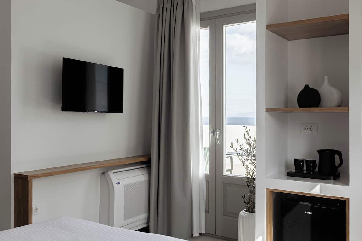 A hotel room with a flat-screen TV mounted on the wall, a bed with a wooden headboard, a radiator, a coffee maker, and a shelf with a vase and cups.