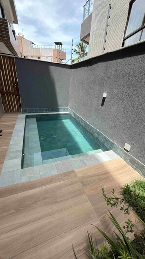 Una piscina rectangular con borde de azulejos, rodeada por una terraza de madera y una pared con textura.