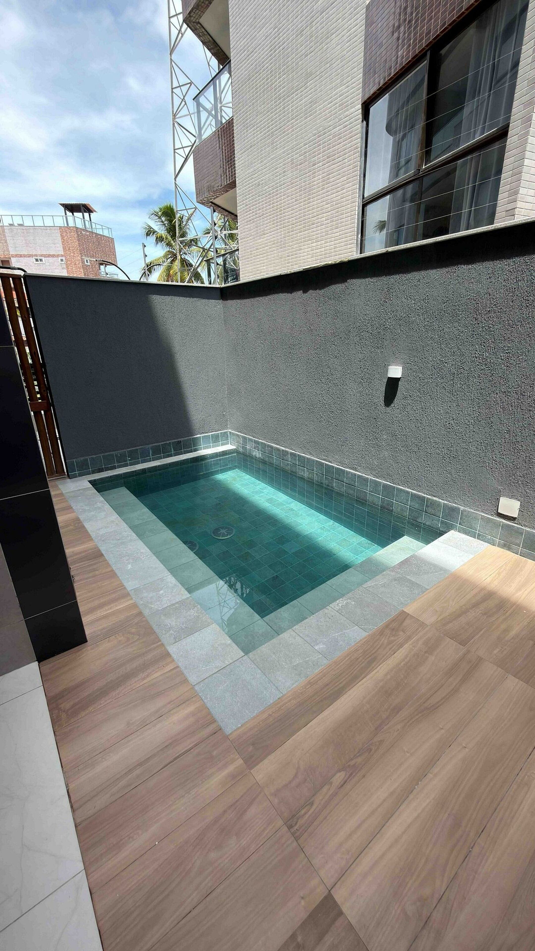 Une piscine rectangulaire dont le bord est recouvert de carreaux, entourée d’un mur gris à la texture particulière et dotée d’un sol en bois.