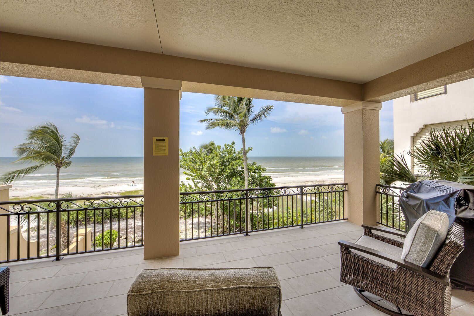A covered balcony with a view of the beach, a wicker chair, and a grill.