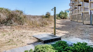 Ein sandiger Pfad führt zu einer Holzterrasse mit einer Bank, umgeben von trockener Vegetation und einem Holzzaun.