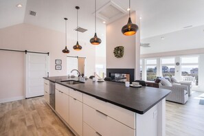 A modern kitchen with a large island, pendant lights, and a view to the living area. - Gorgeous semi-oceanfront home on the 4x4 beaches w/ swim spa & ocean views! (Carova)