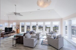 A spacious living room with a large TV, comfortable seating, and a balcony with a view. - Gorgeous semi-oceanfront home on the 4x4 beaches w/ swim spa & ocean views! (Carova)