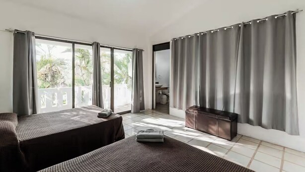 Quarto de hotel com duas camas, um pequeno espaço para sentar, banheiro visível através de uma porta aberta e janelas grandes com cortinas.