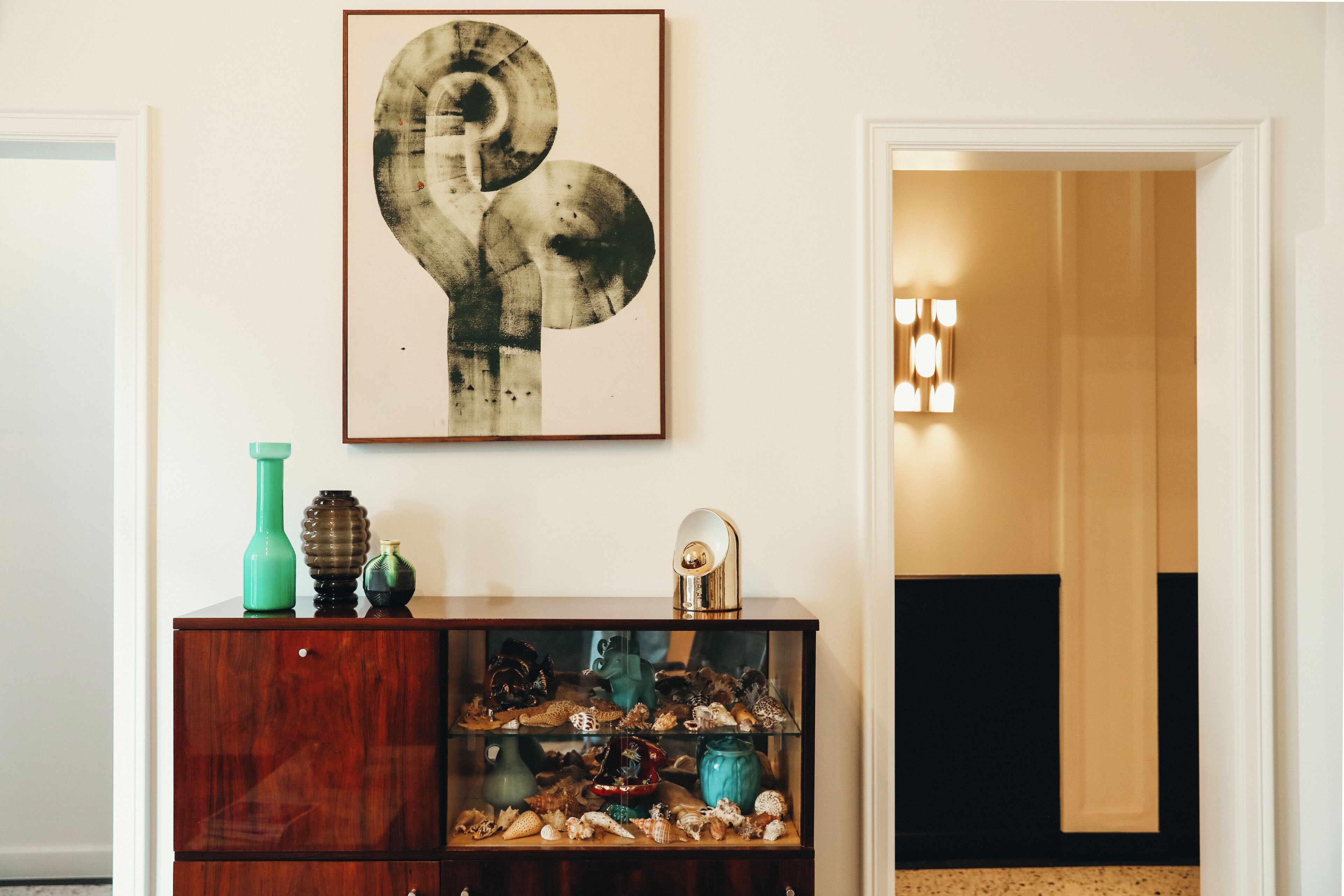 A room with a wooden cabinet displaying seashells and a framed abstract artwork.